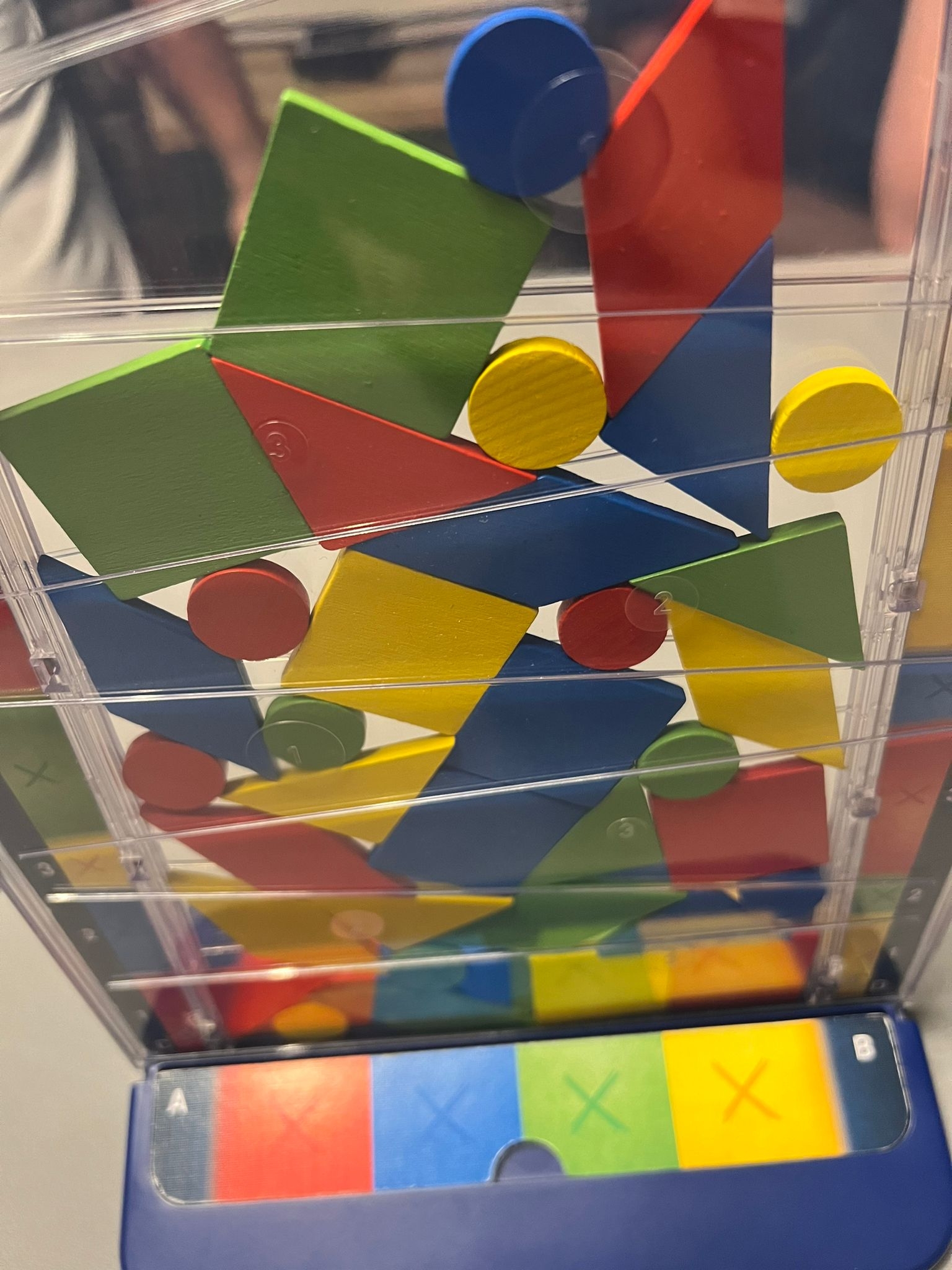 Spiel Drop It. Senkrecht stehender, durchsichtiger Plastikschuber, in den bunte, geometrische Holzteile drinnen sind